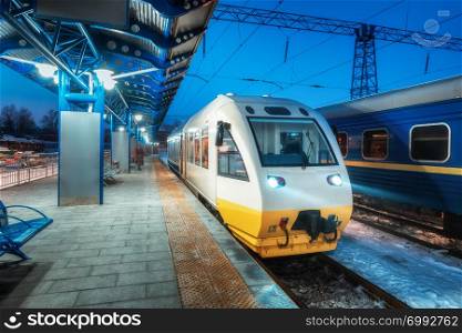 High speed train on the railway station at night in Europe. Urban landscape with modern commuter train on the railway platform with illumination at dusk. Intercity vehicle. Passenger railroad travel