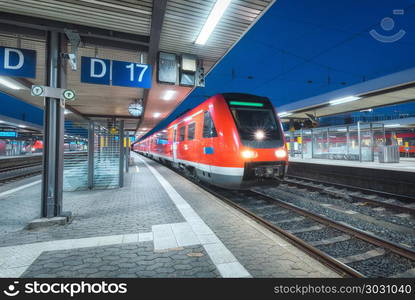 High speed train on the railway station at night in Nuremberg, Germany. Modern intercity train on the railway platform with illumintain. Commuter red train on railroad. Passenger transportation. Dusk. High speed train on the railway station at night