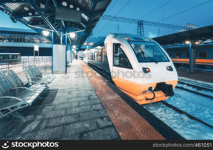 High speed train on the railway station at night in winter. Urban landscape with modern commuter white train on the railway platform with illumination at dusk. Intercity. Passenger railroad travel