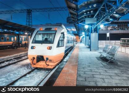High speed train on the railway station at night in winter. Urban landscape with modern commuter train on the railway platform with illumination at dusk. Intercity vehicle. Passenger railroad travel