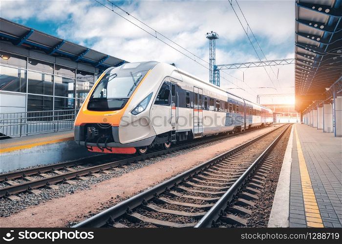 High speed train on the railway station at sunset. Industrial landscape with modern intercity passenger train on the railway platform and blue cloudy sky. Railroad in Europe. Commercial transportation