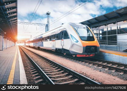 High speed train on the railway station at sunset. Industrial landscape with modern intercity passenger train on the railway platform and blue cloudy sky. Railroad in Europe. Commercial transportation. High speed train on the railway station at sunset