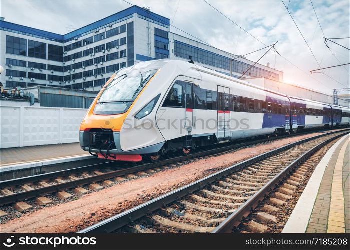 High speed train on the railway station at sunset. Industrial landscape with moving modern intercity passenger train on the railway platform, buildings. Railroad in Europe. Commercial transportation