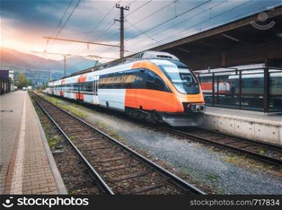 High speed train on the railway station in mountains at sunset in summer. Orange modern commuter train on the railway platform. Industrial landscape with railroad. Passenger transportation. Intercity. High speed train on the railway station in mountains at sunset