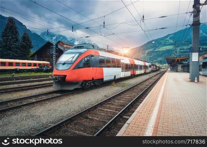 High speed train on the railway station in mountains at sunset in summer. Orange modern commuter train on the railway platform. Industrial landscape with railroad. Passenger transportation. Intercity. High speed train on the railway station in mountains at sunset