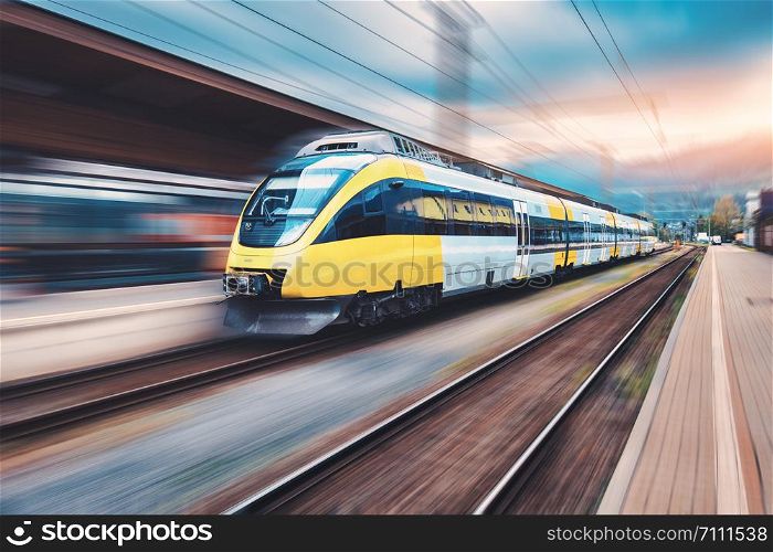 High speed yellow train in motion on the railway station at sunset. Modern intercity passenger train with motion blur effect on the railway platform. Industrial. Railroad and blurred background