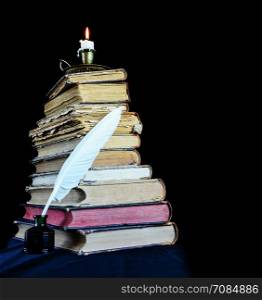 High stack of old books, a burning candle in a candlestick and a white feather in the inkwell
