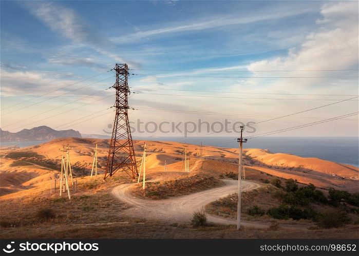 High voltage tower, road, hills with yellow grass in mountains on the background of colorful sky with clouds at sunset. Electricity pylon system in the hill. Summer evening. Industrial landscape