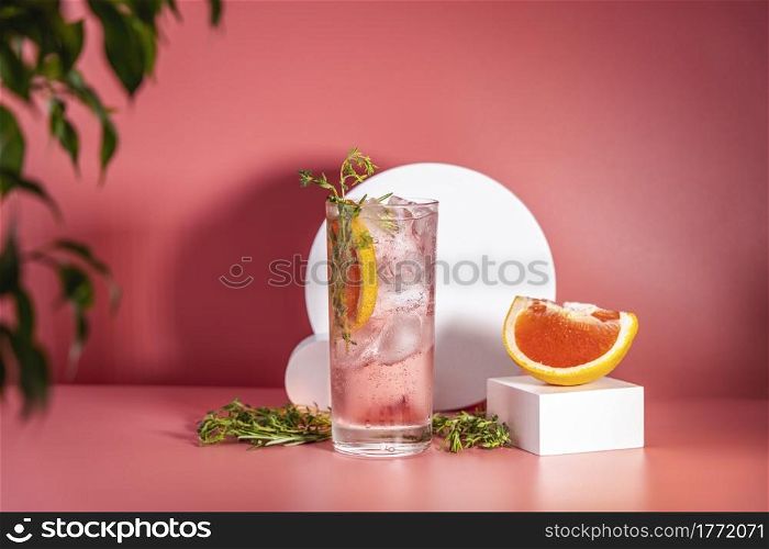 Highball glass of fresh grapefruit cocktail with ice, rosemary and thyme on pink table surface, spring summer art drink food concept.
