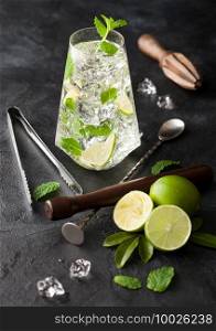 Highball glass of Mojito cocktail with ice cubes,mint and lime on black board with spoon and wooden muddler and fresh limes with ice tongs and squeezer. Best party drink.