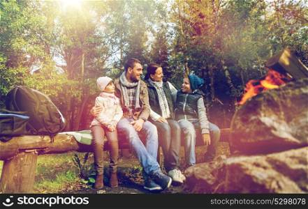 hike, travel, tourism and people concept - happy family sitting on bench and talking at camp near campfire in woods. happy family sitting on bench at camp fire
