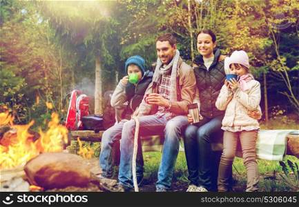hike, travel, tourism and people concept - happy family sitting on bench and drinking hot tea from cups at camp fire in woods. happy family sitting on bench at camp fire