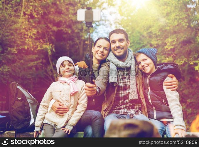 hike, travel, tourism and people concept - happy family sitting on bench and taking picture with smartphone on selfie stick at campfire in woods. family with smartphone taking selfie near campfire
