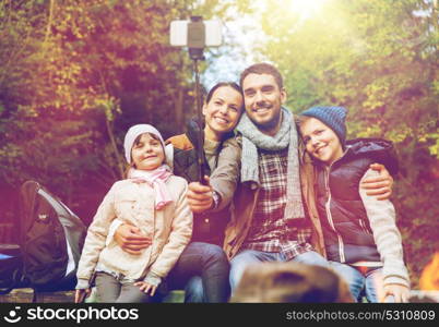 hike, travel, tourism and people concept - happy family sitting on bench and taking picture with smartphone on selfie stick at campfire in woods. family with smartphone taking selfie near campfire