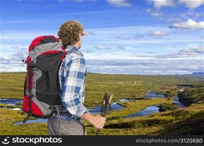 Hiker looking into the distance, upstream a small river, shielding his eyes and holding two walking sticks
