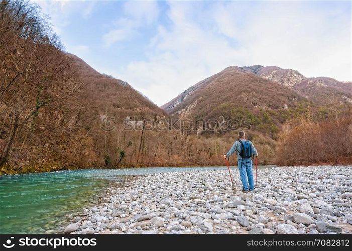Hiker on the bank of a river. Trekking toward mountain. Rambler about 60 years old.