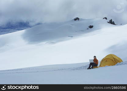 Hiker using laptop outside of tent on snowy mountain peak