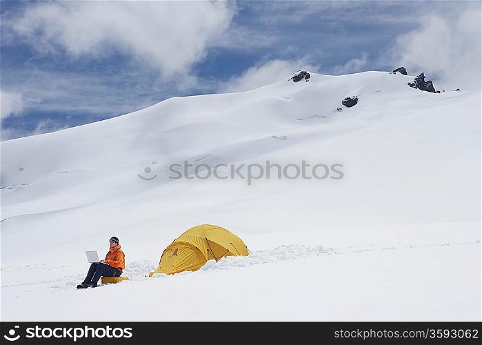 Hiker using laptop outside of tent on snowy mountain peak