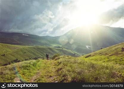 Hiker with a backpack walking through mountain valley, among green hills and alpine meadows; lens flare and sun beams. Lagonaki, Caucasus, Russia