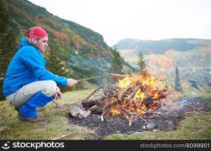 hiking man prepare tasty sausages on campfire in mountains