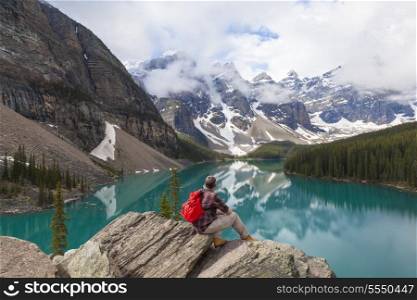 Hiking man sitting down with rucksack backpack standing on tree log by Moraine Lake looking at snow covered Rocky Mountain peaks, Banff National Park, Alberta Canada