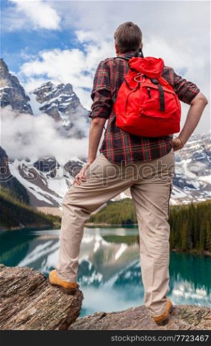 Hiking man with red rucksack backpack standing on a rock overlooking Moraine Lake looking at snow covered Rocky Mountain peaks, Banff National Park, Alberta Canada