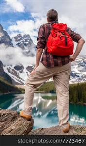 Hiking man with red rucksack backpack standing on a rock overlooking Moraine Lake looking at snow covered Rocky Mountain peaks, Banff National Park, Alberta Canada