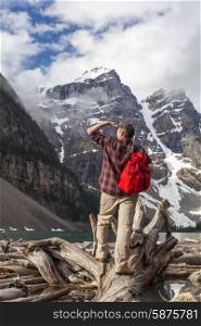 Hiking man with rucsac backpack standing on tree log by Moraine Lake looking at snow covered Rocky Mountain peaks, Banff National Park, Alberta Canada
