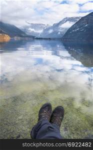 Hiking theme image with the Dachstein mountains and their reflection in the Hallstatter lake water, while a man is relaxing on the shore, with its legs above the water.