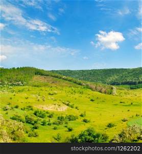 Hilly green fields with trees and shrubs. There are beautiful clouds on the blue sky.
