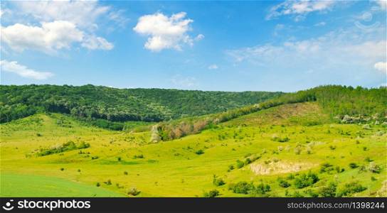 Hilly green fields with trees and shrubs. There are beautiful clouds on the blue sky. Wide photo .
