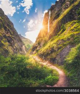 Himalayan mountains covered green grass, high waterfall, beautiful path, green trees, blue sky with yellow sun and clouds in Nepal at sunset. Mountain canyon. Summer travel in Himalayas.Landscape. Mountains covered green grass, high waterfall at sunset