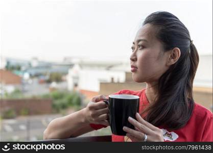 Hip Young Asian Woman Drinking Cup of Coffee on Balcony with Urban Background Scene
