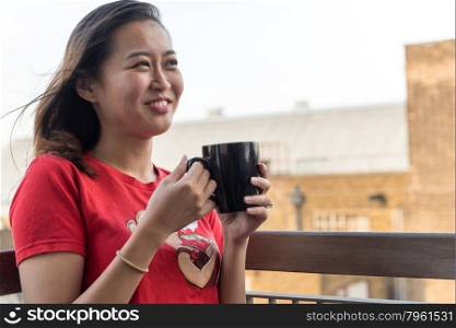 Hip Young Asian Woman Drinking Cup of Coffee on Balcony with Urban Background Scene