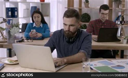 Hipster businessman with beard suffering headache, taking painkillers at workplace while diverse coworker working on background. Sick employee taking medicine in pills while sitting in front of laptop
