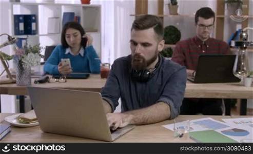 Hipster businessman with beard taking prescription medicine while sitting in front of laptop in office as colleagues working on background. Young freelancer taking drugs from pill bottle at workplace.
