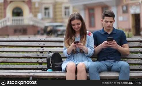Hipster couple sitting on the bench in park, browsing in smartphones and showing mutual disinterest. Concept of relationship apathy, usage of new technology and mobile phone addiction.