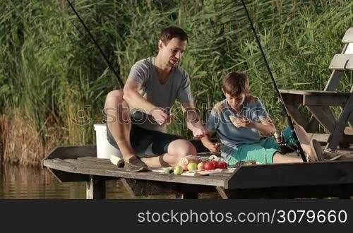 Hipster dad and teenage son having breakfast on wooden pier at the pond while fishing. Father and carefree boy enjoying meal by the lake on sunny summer. Family spending weekend together in countryside. Slow motion.