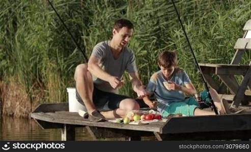 Hipster dad and teenage son having breakfast on wooden pier at the pond while fishing. Father and carefree boy enjoying meal by the lake on sunny summer. Family spending weekend together in countryside. Slow motion.