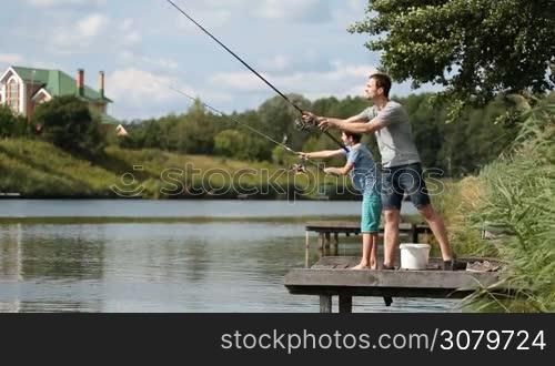 Hipster father and teenage son angling together on wooden pier by the lake on summer day against amazing rural landscape. Positive dad and his cute boy casting fishing rods while spending leisure fishing at freshwater pond. Side view.