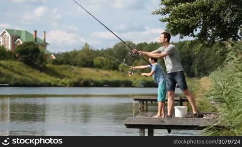 Hipster father and teenage son angling together on wooden pier by the lake on summer day against amazing rural landscape. Positive dad and his cute boy casting fishing rods while spending leisure fishing at freshwater pond. Side view.