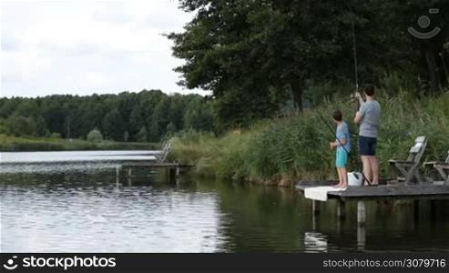 Hipster father standing on wooden pier casting fishing rod on lake and teaching his teenage son how to fish over rural landscape background. Handsome dad and boy spending weekend by freshwater pond fishing.