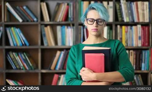 Hipster female student in trendy eyeglasses holding books at library with lovely smile. Portrait of beautiful happy student girl with books smiling to the camera at college library over bookshelves background.