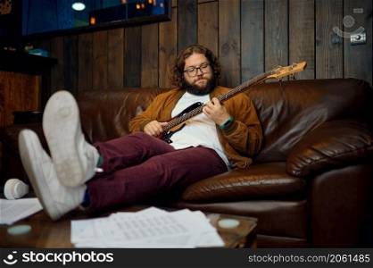 Hipster man playing guitar sitting on couch at home in evening. Creative hobby, rest and relax time. Man playing guitar at home in evening