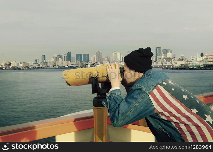 hipster man wearing american flag jeans jacket looking through binocular lens against urban building