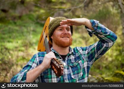 Hipster man with red beard holding a guitar and looking for something in the forest