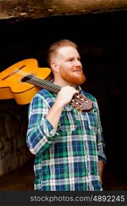 Hipster man with red beard holding a guitar in a rustic house
