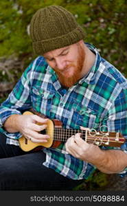 Hipster man with red beard playing a ukulele in the field