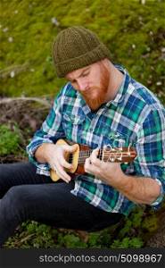 Hipster man with red beard playing a ukulele in the field