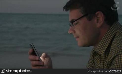Hipster style man in glasses and check shirt using smart phone on the beach in the evening. He texting sms or chatting in social network service. Sea surf in background